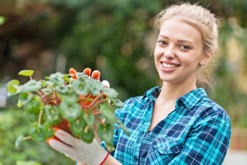 A young woman with blonde hair tied back, wearing a blue and green checked shirt and white gardening gloves, holding a small potted plant with green, slightly variegated leaves and tiny white flowers, in an outdoor garden setting with blurred green foliage in the background. The garden appears well-maintained, with natural grass and a mix of shrubs and trees. This scene exemplifies professional gardening or garden maintenance activities, such as planting or pruning, common in residential outdoor spaces in Bounds Green, London, highlighting the importance of regular garden care provided by services like Lawn Mowing Bounds Green.