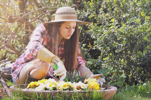 A young woman wearing a wide-brimmed straw hat, a plaid shirt, and gardening gloves is tending to a flower bed in a lush, green outdoor garden space. She is kneeling on the grass, carefully planting or arranging colourful flowers within a circular bed bordered by natural soil and surrounding grass. Behind her, dense foliage and a hedge provide privacy and shade, with sunlight filtering through the leaves, creating a warm and vibrant atmosphere. The garden area features a well-maintained lawn with a variety of plants, including shrubs and small trees, emphasizing a tidy and landscaped yard typical of residential gardens in Bounds Green. This scene showcases regular outdoor maintenance, such as planting and tending flowers, aligning with professional gardening services offered by Lawn Mowing Bounds Green, and highlights the natural beauty of a domestic garden during a clear, sunny day.